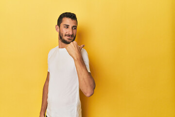 Young Hispanic man on yellow background points with thumb finger away, laughing and carefree.