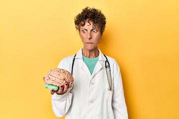 Doctor holding a brain model on yellow studio confused, feels doubtful and unsure. - obrazy, fototapety, plakaty