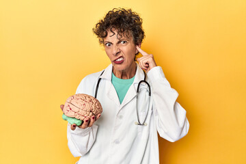 Doctor holding a brain model on yellow studio covering ears with hands.