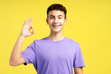 Handsome smiling boy with dental braces wearing casual purple t shirt gesturing, showing ok sign