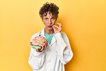 Doctor holding a brain model on yellow studio with fingers on lips keeping a secret.