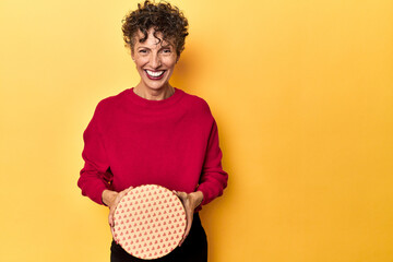 Mature woman holding a heart-adorned box on a yellow studio backdrop