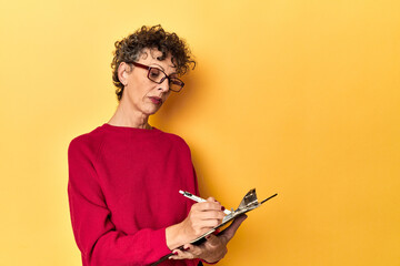 Middle-aged woman jotting down inventory notes on yellow studio background