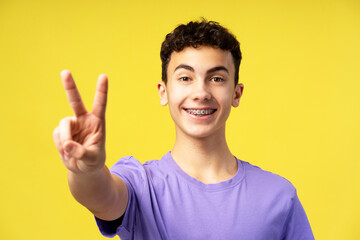 Portrait of happy attractive boy, teenager with braces gesturing, showing peace sign with fingers