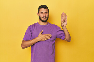 Young Hispanic man on yellow background taking an oath, putting hand on chest.