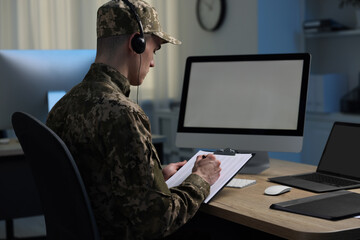 Military service. Soldier with clipboard and headphones working at table in office at night