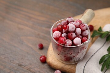 Frozen red cranberries in glass pot and green leaves on wooden table, closeup. Space for text