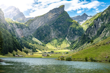 Panoramic view of tourists walking along an alpine lake in a green valley with a mountain peak in...