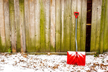 Ready for winter: red snow shovel leans against a wooden back yard fence with leaves and snow in...