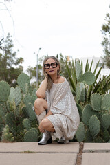 Beauty portrait of a 40-year-old woman, posing in a silver sequin dress and with large-framed sunglasses, where behind there are some cacti