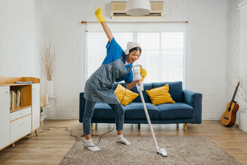 Happy young Asian housewife sings her favorite song with a vacuum cleaner as a microphone while cleaning. Enjoying domestic work dancing in living room a cheerful maid having fun. - obrazy, fototapety, plakaty