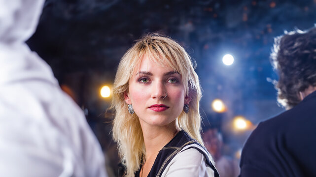 A young woman with blonde hair and a nose ring looks pensively at the camera, lit by ambient stage lights. Generative AI tool used.