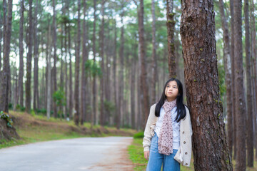 Asian child nature travel or kid girl wearing scarf and sweater stood vacantly leaning against pine tree alone to feel morning lonely in garden forest on holiday vacation at Doi Bo Luang Forest Park