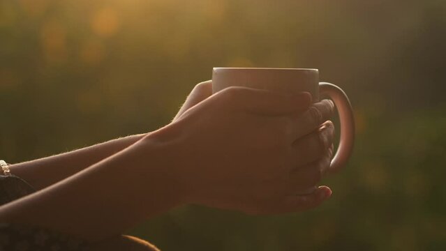 Close-up female hands holding a white ceramic cup of hot coffee or tea to drink at sunrise outdoors. Steam above the mug on blurred nature background. Peaceful morning concept.