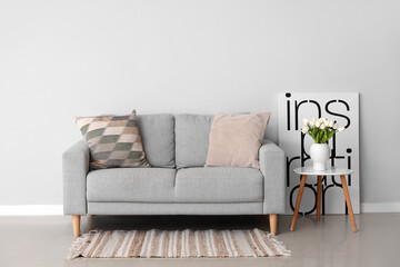 Cozy sofa with cushions and tulip flowers in vase on table near grey wall