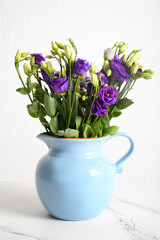 Jug with eustoma flowers on table near white wall