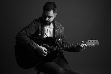 Black and white portrait of a caucasian man playing an acoustic guitar. He is in his 40s and is wearing a jean jacket and a white sweater underneath. The photo was taken in a studio using hard light. 