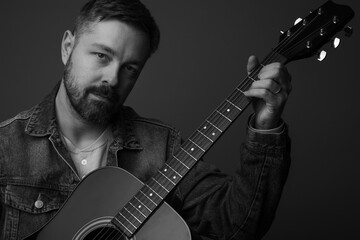 Close up portrait of a caucasian man in his 40s holding and posing with an acoustic guitar. He is wearing a jean jacket and has a beard and hair. He is handsome.