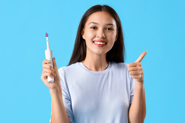 Smiling young Asian woman with electric toothbrush showing thumb-up on blue background