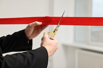 Woman cutting red ribbon with scissors indoors, closeup