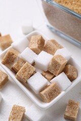 Different sugar cubes in bowl on white table, closeup