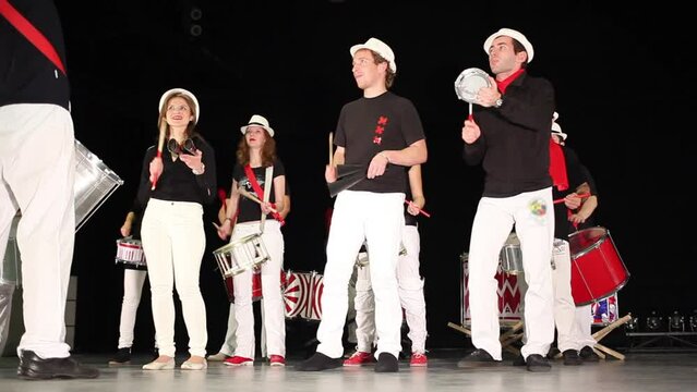 Twelve people in hats play drums in concert hall on stage
