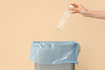 Female hand throwing empty plastic bottle into trash bin near beige wall, closeup