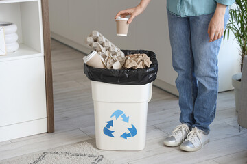 Woman throwing paper cup into full trash bin with recycle logo in kitchen