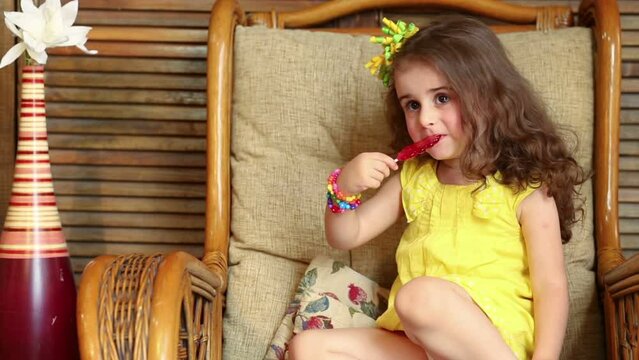 Little curly girl is licking a lollipop and waving ones hand.