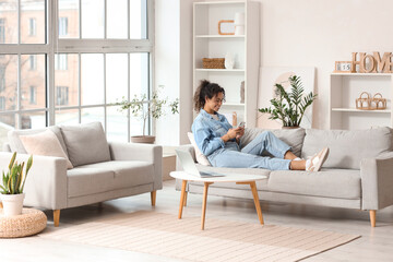 Young African-American woman using mobile phone on sofa at home - obrazy, fototapety, plakaty