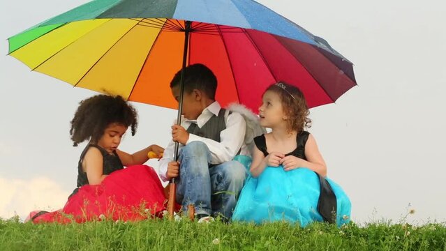 Three bi-racial, mixed race children sit on grass under umbrella during rain.