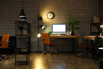 Interior of dark office with desks, shelf unit and glowing lamps at night