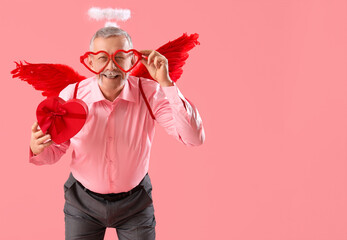 Mature man dressed as Cupid with heart-shaped eyeglasses and gift on pink background. Valentine's...