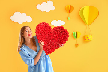 Young woman with heart near decorated yellow wall. Valentine's Day celebration