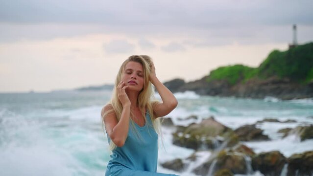 Blonde woman in blue dress enjoys serene sea breeze at rocky coast. Elegant lady hair, contemplates ocean waves. Relaxation, feminine beauty tropical beach. Natural allure outdoors.