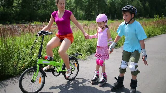 Boy and girl roller-skate and mother rides bike and hold hands