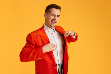 stylish winking young man in orange jacket and striped pants on vibrant yellow background.