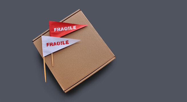 Small paper flags with the warning 'Fragile' as a label and empty cardboard box on monochrome gray background. The concept of shipping fragile products. Copy space