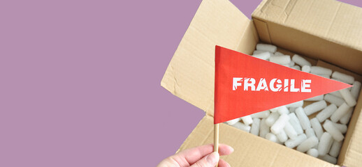 Opened cardboard packaging box with white polystyrene packaging chips. Tiny red paper flag in hand with the warning 'Fragile' as a label. The concept of shipping fragile products