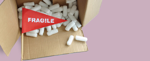 An overturned open cardboard packaging box with fallen white polystyrene packaging chips. Tiny paper flag with the warning 'Fragile' as a label. The concept of shipping fragile products