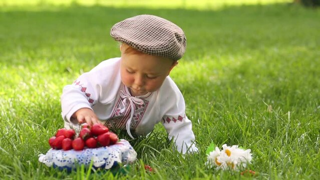 Cute baby in folk costume crawls on grass touches strawberries