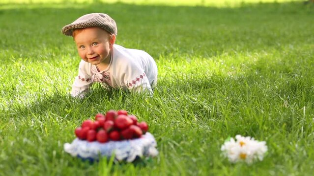 Cute baby in folk costume crawls on grass to red strawberries