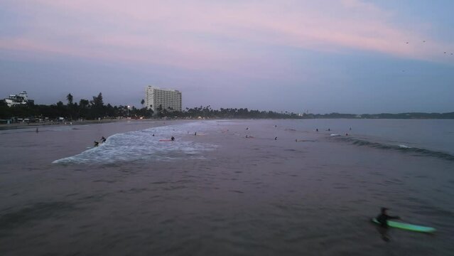 Surfers catch waves at dusk in crowded sea. Beginner surfers paddle out as sunset colors sky. Aerial view shows shoreline, waves breaking, surfing lessons at tropical beach with hotels.