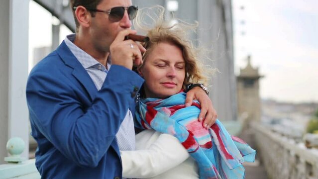 Woman hugs man smoking cigar and strokes his cheek on bridge