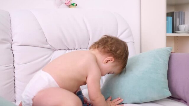 Little kid in diaper plays with toys on white sofa with pillows