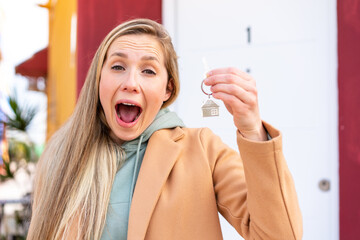Young blonde woman holding home keys at outdoors with surprise and shocked facial expression