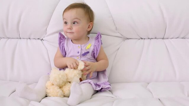 Little baby in dress with toy sits on white sofa at home