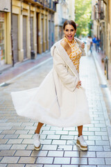 Naklejka premium Content stylish woman in fashionable dress and coat standing on street against buildings