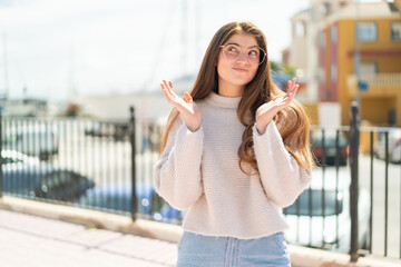 Young pretty caucasian woman with glasses at outdoors making doubts gesture