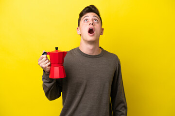 Young Brazilian man holding coffee pot isolated on yellow background looking up and with surprised...